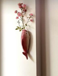 a fish shaped vase with pink flowers hanging on the wall next to a white wall