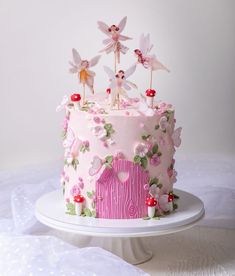 a pink cake decorated with fairy figurines and flowers on a white tablecloth