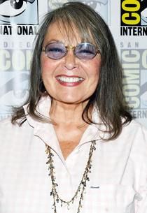 an older woman wearing sunglasses and smiling for the camera at a press room event in front of a comic panel