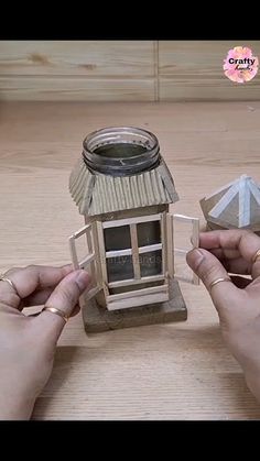 two hands are holding small paper houses on a wooden table, one has a candle in it and the other is made out of wood