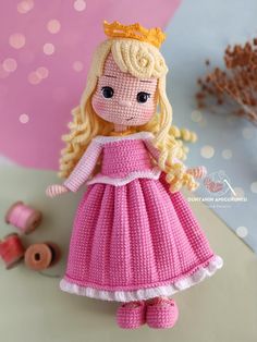 a crocheted doll wearing a pink dress and tiara, standing next to spools of thread