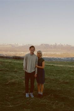 a man and woman standing next to each other in a field with mountains in the background