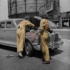 two women working on an old car in front of a large water tank, 1950's or 1960s's