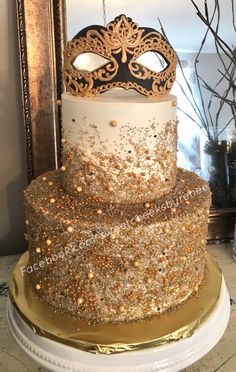 a three tiered cake decorated with gold and white frosting, topped with a masquerade mask