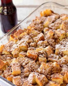 a casserole dish with bread and cinnamon toppings in a glass baking dish next to a bottle of syrup