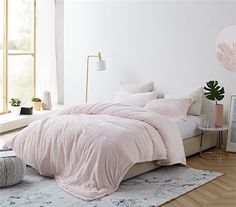 a bed with pink comforter and pillows in a white room next to a window