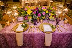 the table is set with purple and yellow flowers, gold place settings, and silverware