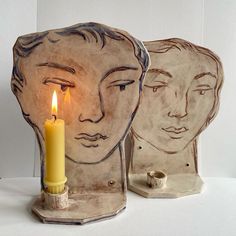 a candle that is sitting in front of two faces on a table with a white wall behind it