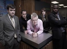 a group of people standing around a table in an office setting with one man leaning on the counter