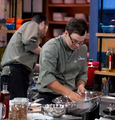 two men in the kitchen preparing food on top of a table with utensils