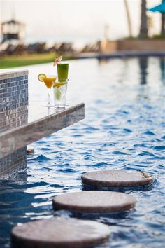 two cocktails are sitting on the edge of a pool with stepping stones in it