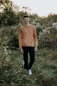 a man standing in tall grass with his hands in his pockets and smiling at the camera