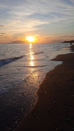 the sun is setting over the water at the beach with people walking on it and in the distance