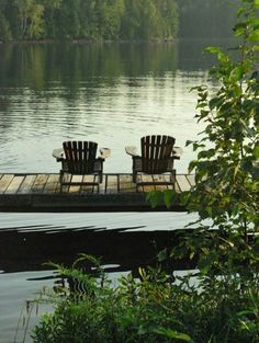 two empty chairs sitting on a dock in the middle of a lake with trees around them