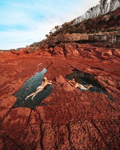 two people are swimming in the water on some red rocks and dirt, with one person laying down