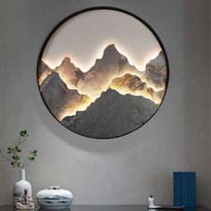 a round mirror with mountains in the background on a wall above a table and vase