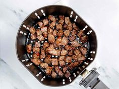 meat cooking in a frying pan on top of a marble counter with tongs