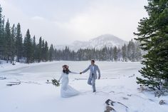 a bride and groom are holding hands in the snow near some trees, with mountains in the background