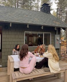 three women sitting at a picnic table with their dogs on the back porch in front of a cabin