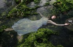 a woman laying on the ground covered in green moss next to a river with water flowing over her