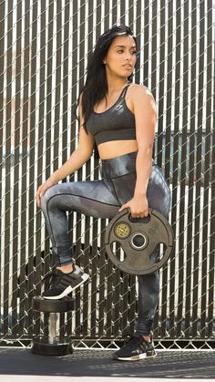 a woman holding a barbell in front of a metal fence while wearing leggings