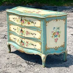 an old dresser painted with flowers and birds on it's sides is sitting in the gravel