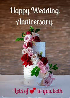 a white wedding cake with red and pink flowers on it, says happy wedding anniversary lots of to you both