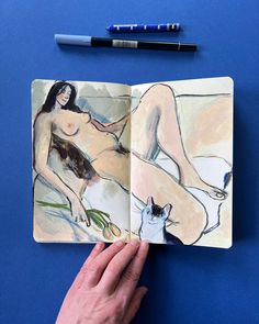 a hand holding an open book with a drawing of a naked woman and a cat