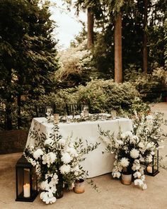 a table with flowers and candles on it in the middle of some bushes, surrounded by trees