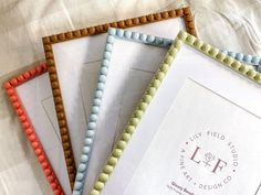 four different colored beaded frames sitting on top of a white bed sheet with the words lilly field studio written below them