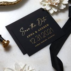 a black and gold save the date card next to some white flowers on a table