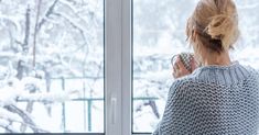 a woman standing in front of a window holding a cup and looking out the window