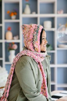 a woman wearing a crocheted hat and scarf sitting at a table in front of a bookcase