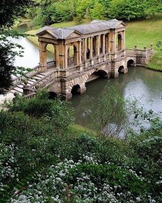 an old bridge over a small pond in the middle of a field with trees and flowers