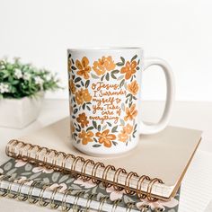 a coffee mug sitting on top of a notebook next to a pen and flower pot