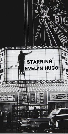 a black and white photo of a man standing on a ladder in front of a movie theater