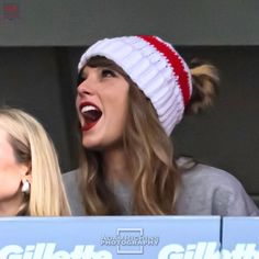 two women laughing while sitting in the stands at a sporting event, one wearing a red and white knitted hat