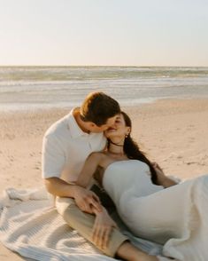 a man and woman sitting on top of a beach next to the ocean with their arms around each other