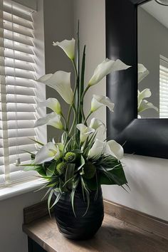 a black vase with white flowers sitting on a wooden table in front of a mirror