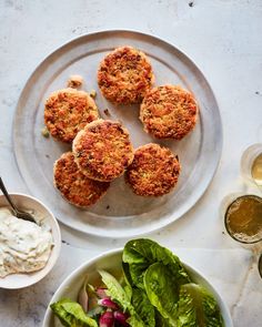 four crab cakes on a plate next to a bowl of salad and two cups of dressing