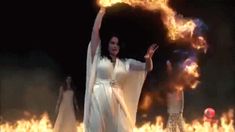 two women dressed in white are dancing with fire and flames behind them, one is holding her arms up