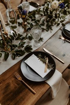 the table is set with black plates and silverware, greenery, and menu cards