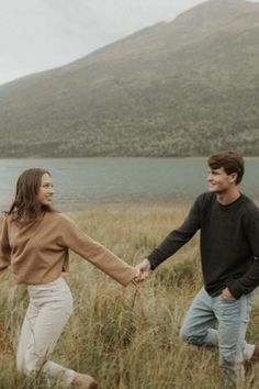 a man and woman holding hands while walking through tall grass near a body of water