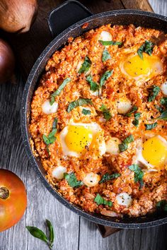 an egg dish in a skillet on a wooden table with tomatoes and onions next to it