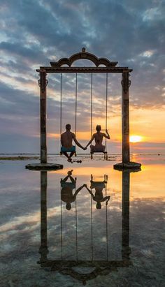 two people holding hands while sitting on swings in front of the ocean at sunset or sunrise