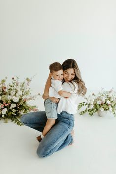 a mother holding her son while he is sitting on the floor in front of flowers