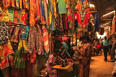 people are shopping in an open market with colorful fabrics and scarves hanging from the ceiling