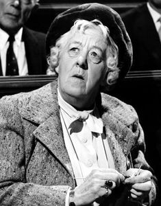 an old woman in a suit and hat sitting at a table with other men behind her