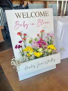 a welcome sign with flowers in it sitting next to a white box filled with baby's breath