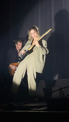 a woman holding a guitar in her right hand while standing on stage with another man behind her